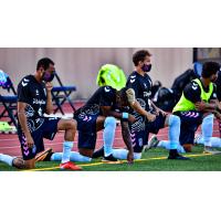 Forward Madison FC players kneel  on the sideline